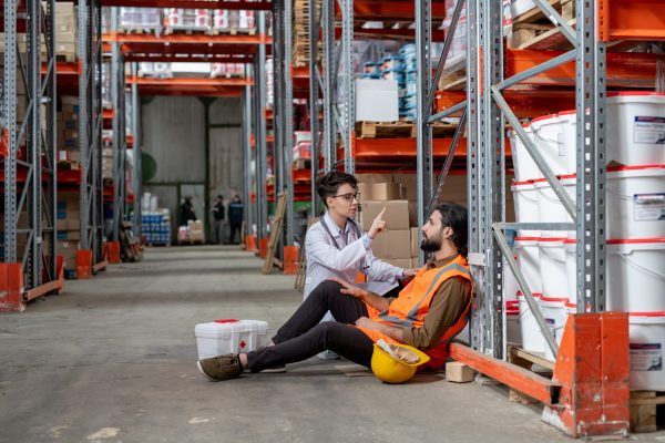 Young female giving first aid to warehouse worker with headache